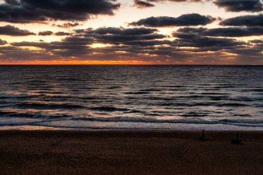 Bulgaristan 'ın Obzor kentindeki Karadeniz üzerinde katmanlı bulutlar, yumuşak turuncu ufuk ışığı ve boş kumlu plajlı dramatik bir gün doğumu. Dalgalar ve kıyı havasıyla huzurlu bir deniz manzarası..