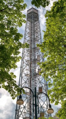 Milano 'daki Parco Sempione' da bulunan metal gözlem kulesi Torre Branca, aşağıdan yakalanmış, taze yeşil ağaç yaprakları ve parlak gökyüzü ile çerçevelenmiş, çelik yapısı ve yüksekliğini gösteriyor..