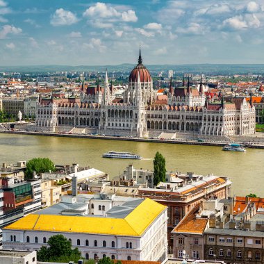 Budapeşte 'deki Tuna Nehri boyunca uzanan Macar Parlamentosu binasının panoramik gündüz manzarası, açık bir gökyüzü altında tarihi mimari, şehir manzarası, çatılar ve turistik tekneleri gösteriyor..