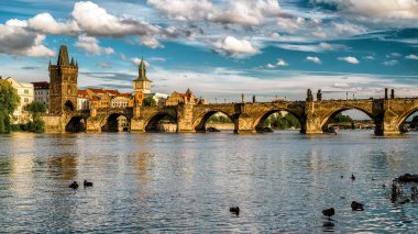 Prag 'daki Charles Köprüsü' nün tarihi kuleleri ve kemerleriyle Vltava Nehri 'ni aydınlık gün ışığı ve dramatik bulutlar altında manzaralı şehir manzarası altında genişletiyor..