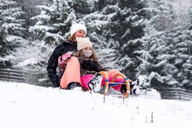 Anne ve çocuk taze kar altında hızla kayıyorlar. Dinamik kış eylemi, açık hava eğlencesi, aile aktivitesi ve orman manzarasında soğuk hava eğlencesi..