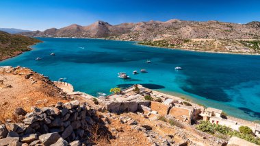 Spinalonga adasının yüksek açılı görüntüsü turkuaz denizi, demirlemiş tekneleri, Girit kıyı şeridinin taş kalıntılarını ve kurak tepelerini açık mavi gökyüzü altında gösteriyor..