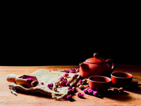 Çaydanlık ve çay fincanı seti. Ahşap masa ve siyah arka plan üzerinde gül çayı yaprakları.