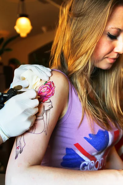 Tatuador que muestra el proceso de hacer un tatuaje en la joven hermosa mujer hipster con la mano de pelo rubio. Diseño del tatuaje en la forma pin-up chica con cabeza de flor — Foto de Stock