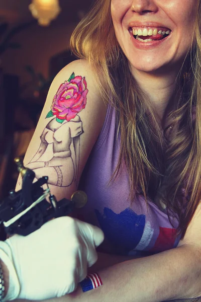 Tatuador que muestra el proceso de hacer un tatuaje en la joven hermosa mujer hipster con la mano de pelo rubio. Diseño del tatuaje en la forma pin-up chica con cabeza de flor — Foto de Stock