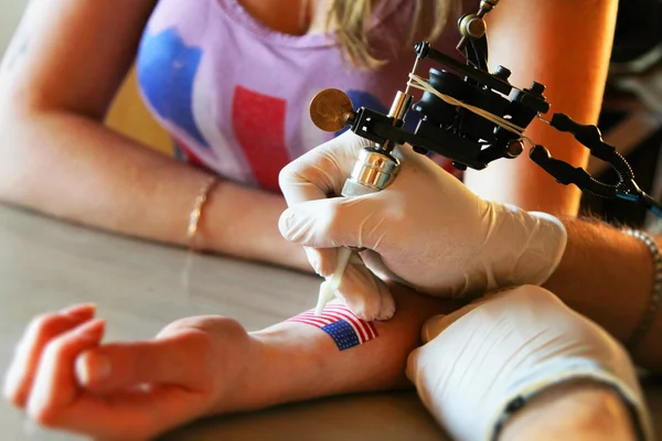 Tattooer showing process of making a tattoo on young beautiful hipster woman with blonde hair hand. Tattoo design in the form pin-up girl with flower head — Stockfoto