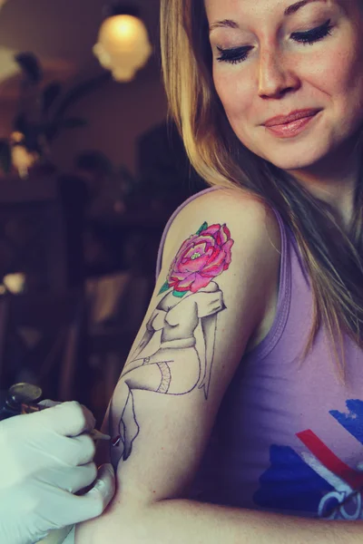 Tatuador que muestra el proceso de hacer un tatuaje en la joven hermosa mujer hipster con la mano de pelo rubio. Diseño del tatuaje en la forma pin-up chica con cabeza de flor — Foto de Stock