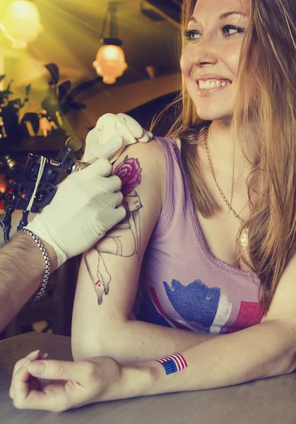 Tattooer showing process of making a tattoo on young beautiful hipster woman with blonde curly hair . Tattoo design in the form of rose — Stock Photo, Image