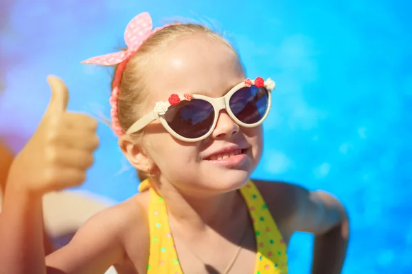Ragazzina che mostra i pollici in su sullo sfondo della piscina. Felice sorridente Bambino indossa occhiali da sole, godendo di piscina e divertirsi. Viaggio (vacanza), concetto di avventura. Da vicino, all'aperto . — Foto Stock