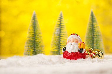 Noel Baba 'nın seramik figürü ve Noel topları karların arasında soyut sarı arka planda Noel ağaçları arasında..