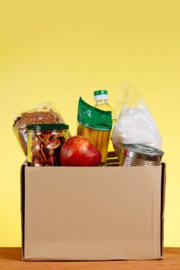 Yiyecek bağışı konsepti. Sarı arka planda bağışlanacak yiyeceklerin olduğu bağış kutusu. Coronavirus salgınının bağlamında yaşlılara yardım. Dikey çekim