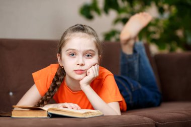 Küçük tatlı ve mutlu kız 9 ya da 10 yaşında, elinde kitapla oturma odasındaki kanepeye uzanmış. Çocuk Eğitim ve Yaşam Tarzı Konsepti.