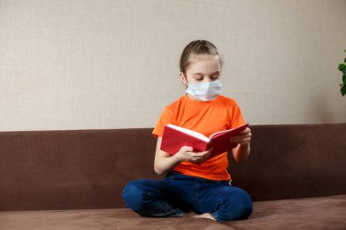 Tıbbi maskeli küçük kız evde kendini izole ediyor ve kitap okuyor.