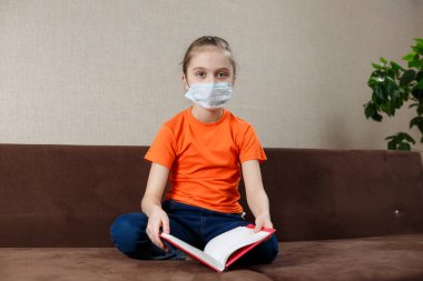 Tıbbi maskeli küçük kız evde kendini izole ediyor ve kitap okuyor.