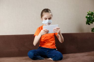 Evde Karantina. Maskeli küçük kız tablet bilgisayar kullanıyor. İnternette film seyrediyor.