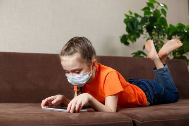 Maskeli küçük kız kanepede uzanıyor ve tablet bilgisayar kullanıyor. Evde yalnızlık