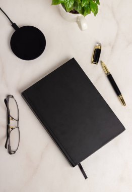 Kablosuz şarj aleti, gözlüğü, siyah not defteri ve mermer arka planda kalemi olan işyeri. 