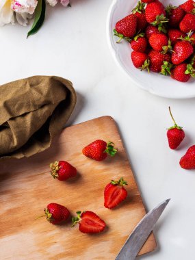 Taze olgun çilekler ve beyaz mermer masadaki tahta kesme tahtasının üzerinde beyaz bir porselen kasede..