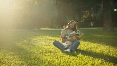Çimenlerin üzerinde bağdaş kurmuş bir kadın altın ışıkta Corgi 'sini tutuyor. Sıcak bir tarlada huzurlu bir yakınlık yakaladığımız an.