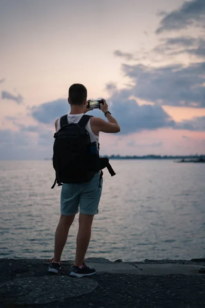 A man photographs the sea and sunset. A man with a backpack on the beach takes photos of the sea at sunset on a smartphone