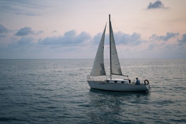 Gün batımında denizde yüzen bir tekne. Beyaz yelkenli gemi bulutların ve günbatımının arka planında.