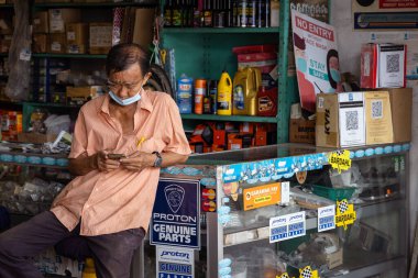 Kuching, Sarawak, Malezya - 15 Nisan 2025: Asyalı oto tamircisi yedek parça dükkanında cep telefonu kullanarak yüz maskesi takıyor