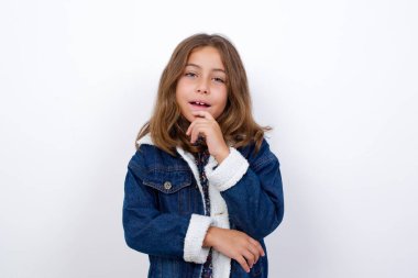 İyimser küçük beyaz kız, güzel mavi gözlü, yalıtılmış beyaz arka plan üzerinde duran kot ceketli elleri kısmen çapraz ve elleri çenenin altında, zevkle kameraya bakıyor. Mutlu duygu konsepti.