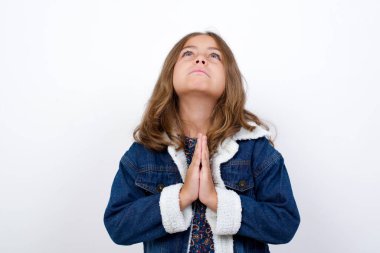 Güzel mavi gözlü beyaz ceketli küçük beyaz bir kız izole edilmiş beyaz arka planda duruyor umut dolu bir ifadeyle birlikte çok duygusal ve endişeli bir şekilde dua ediyor. Lütfen Tanrım.