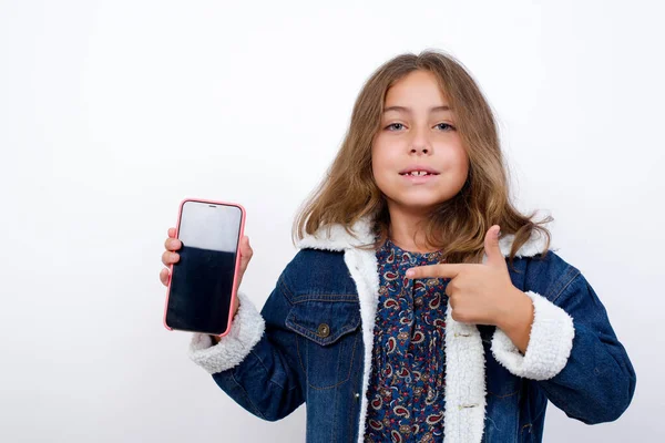 独立した白い背景の上にデニムジャケットを着た美しい青い目をした小さな白人の女の子を笑っています コピースペースをモックアップします 空白の画面を持つ携帯電話上のインデックス指を指す — ストック写真