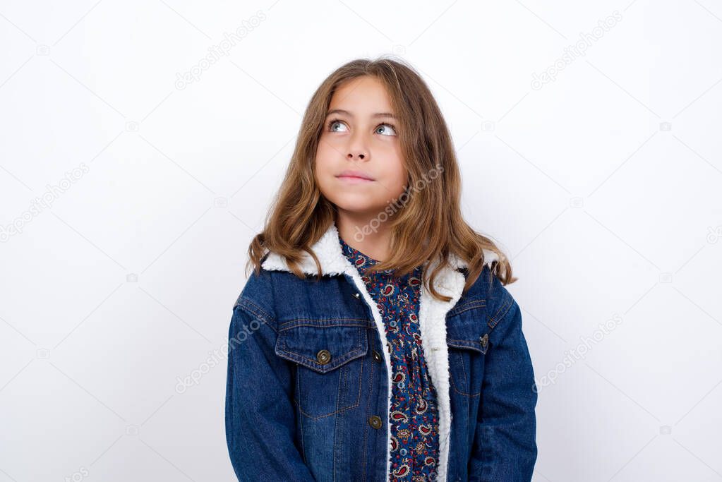 Little caucasian girl with beautiful blue eyes wearing denim jacket standing over isolated white background looking aside into empty space thoughtful 