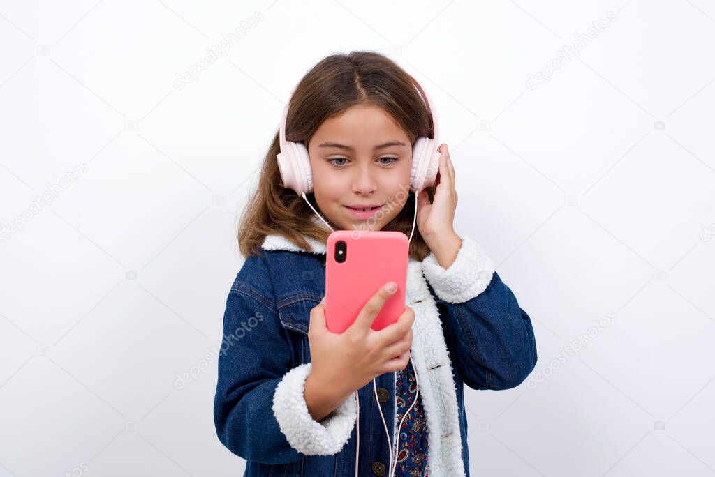 Happy Little caucasian girl with beautiful blue eyes wearing denim jacket standing over isolated white background feels good while focused in screen of smartphone. People, technology, lifestyle