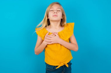 Küçük kız elleri göğsünde, gözleri kapalı gülümsüyor ve yüzünde minnet ifadesi var. Sağlık kavramı.