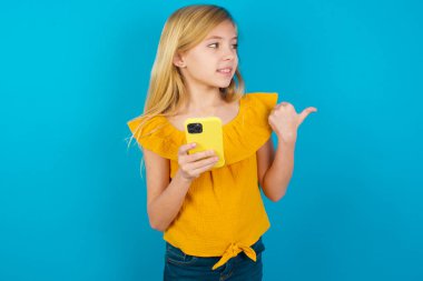 Küçük kız akıllı telefonuyla mesaj atıyor ve gülümseyen yüzüyle baş parmağıyla işaret ediyor.