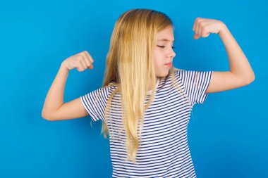 Mavi duvara karşı çizgili gömlek giyen beyaz kız, gururla gülümseyen kol kaslarını gösteriyor. Fitness konsepti