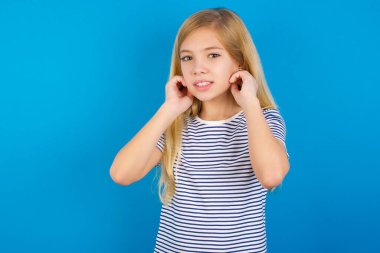 Kafkasyalı kız, yüksek sesli müzik sesi için mavi duvara çizgili gömlek giyip kulakları parmaklarıyla kapatıyor. Sağır kavramı.