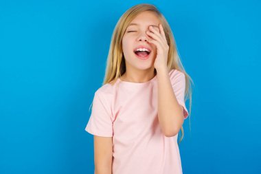 Caucasian kid girl wearing pink shirt against blue wall makes face palm and smiles broadly, giggles positively hears funny joke poses clipart