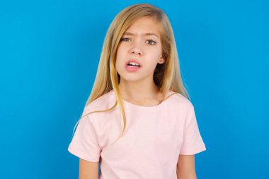 Tatminsiz beyaz kız portresi pembe gömlekli mavi duvara karşı sırıtış, dudaklar, dudaklar ve kameraya rahatsız edici bakışlar, tatsız bir şey duyma hoşnutsuzluğu