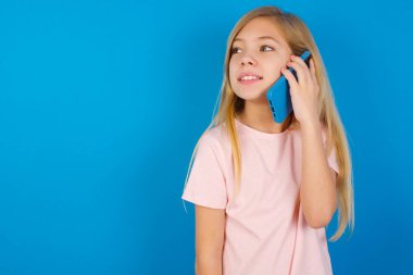 Mavi duvara karşı pembe tişört giyen mutlu beyaz kız güzel bir telefon görüşmesi yapıyor ve yan bakıyor, güzel bir havası var ve cep telefonuyla konuşurken pozitif gülümsüyor.