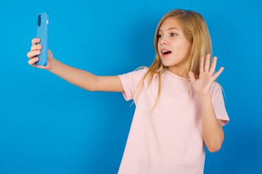 Mavi duvara karşı pembe tişört giyen beyaz bir kız modern cep telefonunu tutuyor ve merhaba jesti olarak video dalgaları yapıyor. İnsanlar modern teknoloji kavramı