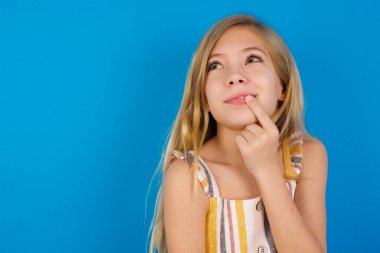 Mavi duvara karşı elbise giyen güzel, hayalperest beyaz kız parmaklarını dudaklarına yakın tutuyor..