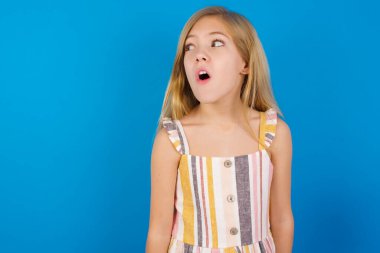 Shocked Caucasian kid girl wearing dress against blue wall look empty space with open mouth screaming: Oh My God! I can't believe this. clipart