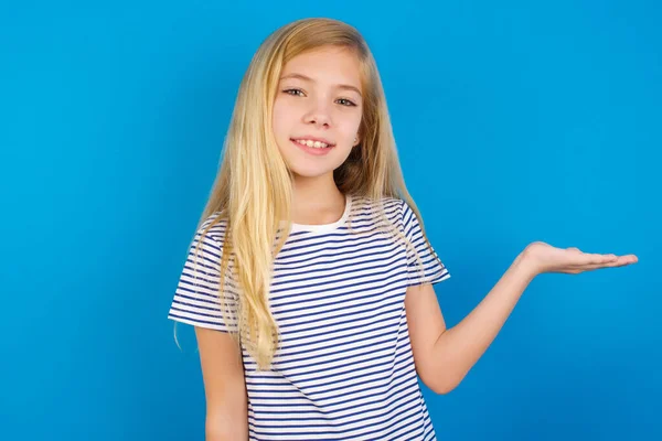 Caucasian girl wearing striped shirt against blue wall, smiling cheerful presenting and pointing with palm of hand looking at the camera