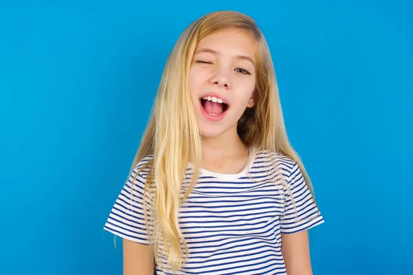 白人の少女は青い壁に縞模様のシャツを着て カメラを見てウィンク 陽気で幸せ — ストック写真