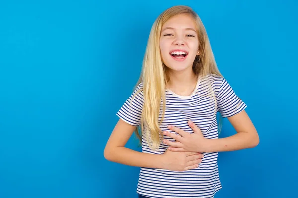 Caucasian girl wearing striped shirt against blue wall, smiling and laughing hard out loud because funny crazy joke with hands on body.
