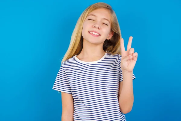 青い壁に縞模様のシャツを着た白人の女の子は カメラで勝利のサインを行うと 幸せな顔をしてウィンク笑顔 第二番 — ストック写真