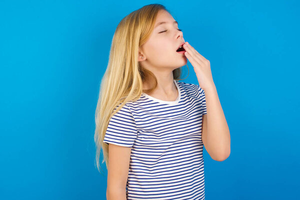 Caucasian girl wearing striped shirt against blue wall being tired and yawning 