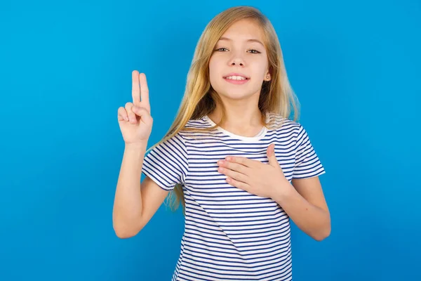 Caucasian girl wearing striped shirt against blue wall smiling swearing with hand on chest and fingers up, making a loyalty promise oath.