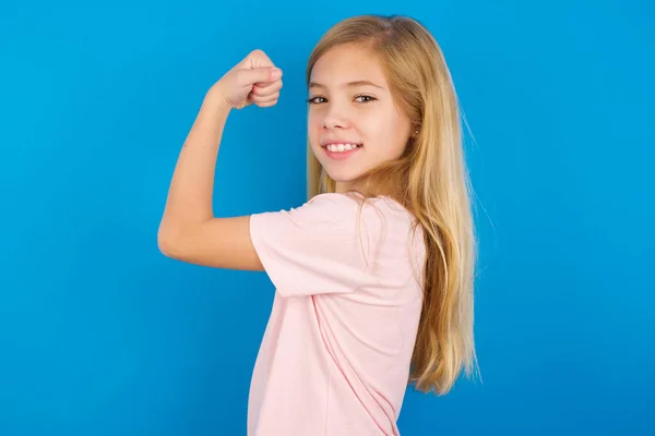 パワフルな陽気な白人の女の子の肖像画ピンクのTシャツを身に着けています青い壁に対して筋肉を示す — ストック写真