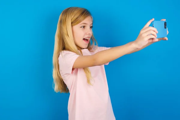 白种人小女孩穿着粉色衬衫 靠着蓝色的墙 带着自拍的照片在社交媒体上发布 或者和朋友们一起打一个视频电话 — 图库照片