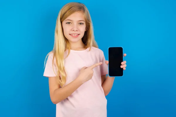 青い壁にピンクのシャツを着た白人の子供の笑顔コピースペースをモックアップします 空白の画面を持つ携帯電話上のインデックス指を指す — ストック写真